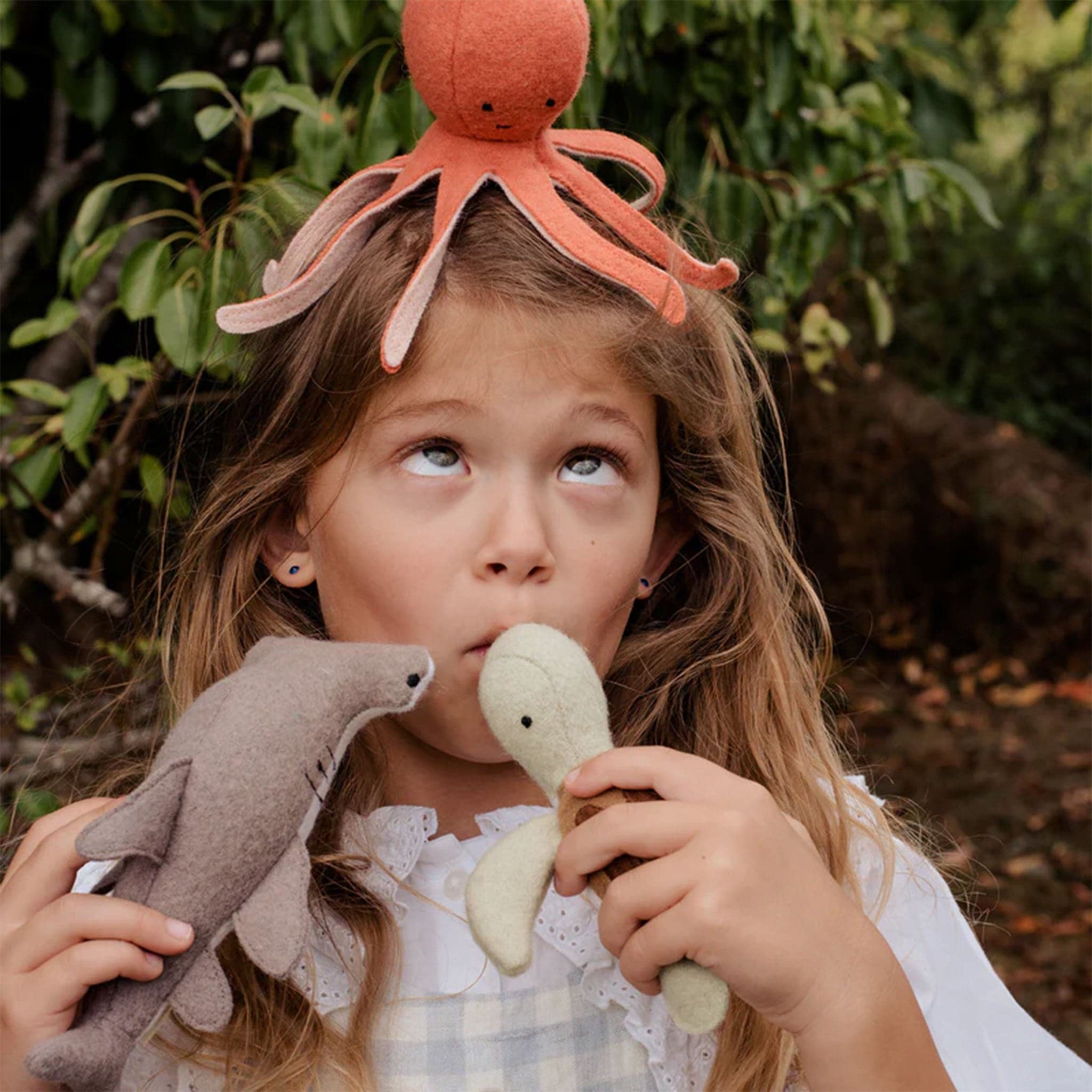 A little girl holding the three marine inspired stuffed toys, that includes an orangey red octopus, a great hammerhead shark and a green and brown sea turtle.