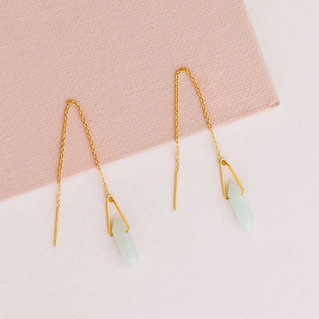 Pair of gold plated chain earrings. Each earring has a soft blue amazonite stone positioned between a gold loop. Pair lays against a soft pink textured box and white counter.