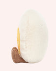 Side view of A soft stuffed animal in the shape of an egg with yellow yoke face and blushing smiley face, and brown floppy legs.