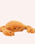 In front of a cream background is a stuffed orange crab laying on its stomach. It has two large claws in the front and two little claws in the back. On its face are two black eyes and a little black smile. He has two orange antennas above his eyes.