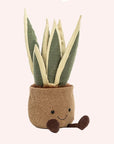 A soft suede stuffed animal in the shape of a potted snake plant, with spikey dark and light green leaves, and brown textured pot with smiley face and dark brown floppy legs.