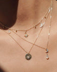 Close up of three layered necklaces being worn. One is a gold choker with star, moon, and white gem charms. One necklace is a long gold necklace with silver gem moon with white gem drop. The adenna necklace is the third necklace. Long gold chain and circle pendant with crystal gemmed star charm.