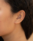 A pair of tiny gold daisy shaped stud earrings with a straight gold post worn here on a model's ear.