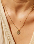 This picture is a close up of a woman’s neck. She is wearing a tan blouse. Around her neck is a gold chain with a gold circle pendent on the end. In the middle of the pendent is a gold rose.