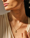 This picture is a close up of a woman’s neck. She is wearing a tan blouse. Around her neck is a gold chain with a gold circle pendent on the end. In the middle of the pendent is a gold rose.