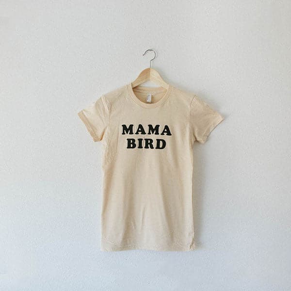 A cream cotton t-shirt with black letters across the from that read, "Mama Bird" hung on a hanger in front of a white background.