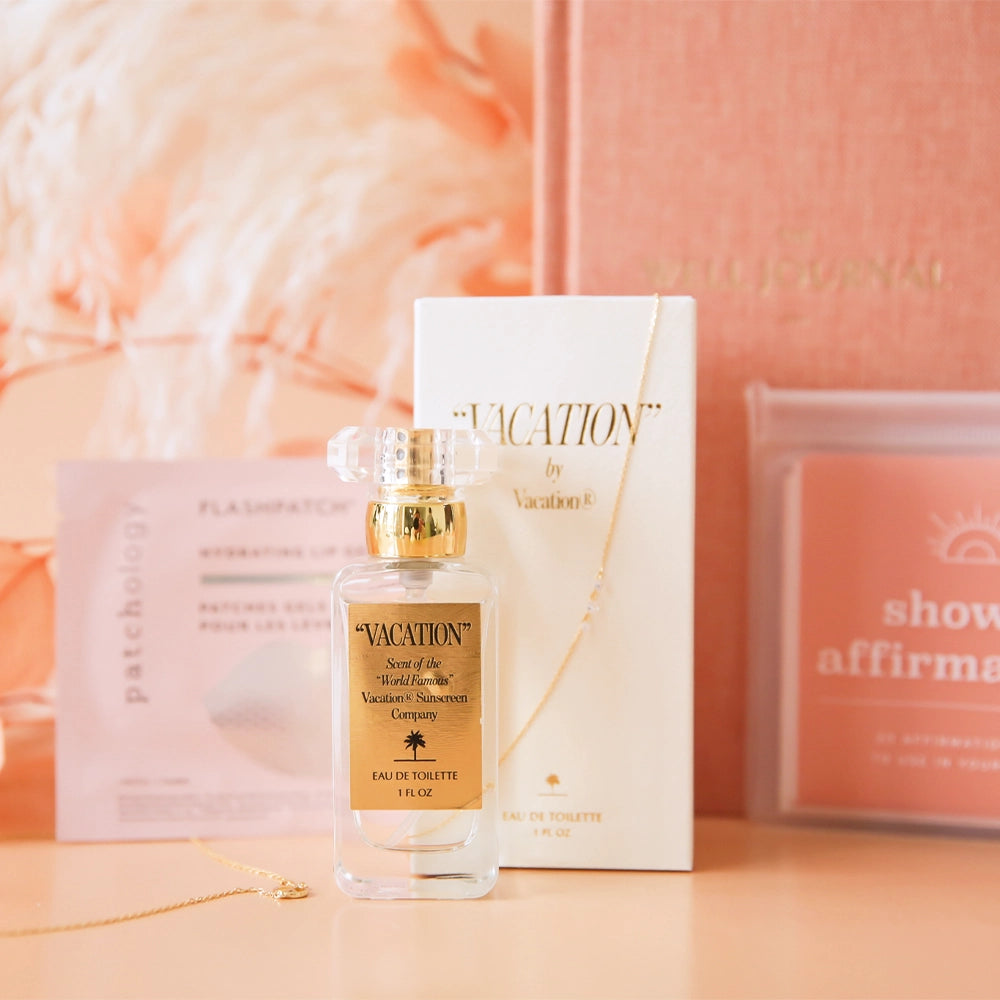 A clear glass bottle of perfume with gold detailing and a gold label that reads, &quot;Vacation scent of the world famous Vacation Sunscreen&quot; in front of an array of pink and white dried florals and products. 