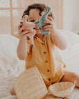 Photo of a child in a golden yellow button up top playing with two small felt dinosaur stuffed animals. Child has brown curly hair and is smiling in between the two dinosaurs. The child is sitting on a cozy bed with white linens and has a small wicker basket in front of them.