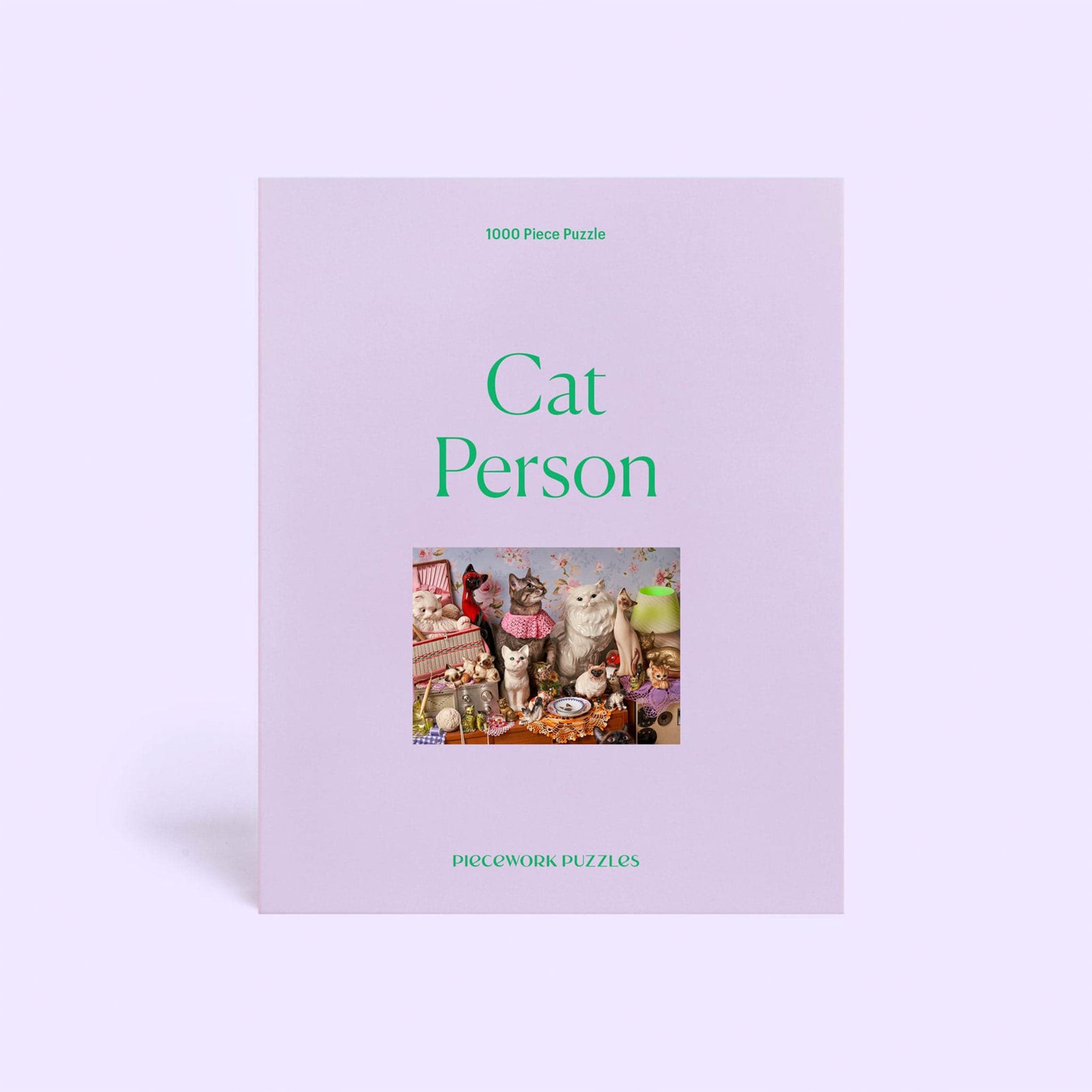 On a lavender background is a 1,000 piece puzzle box featuring the photo of the finished puzzle in the center along with green letters that read, &quot;Cat Person Piecework Puzzles&quot;.