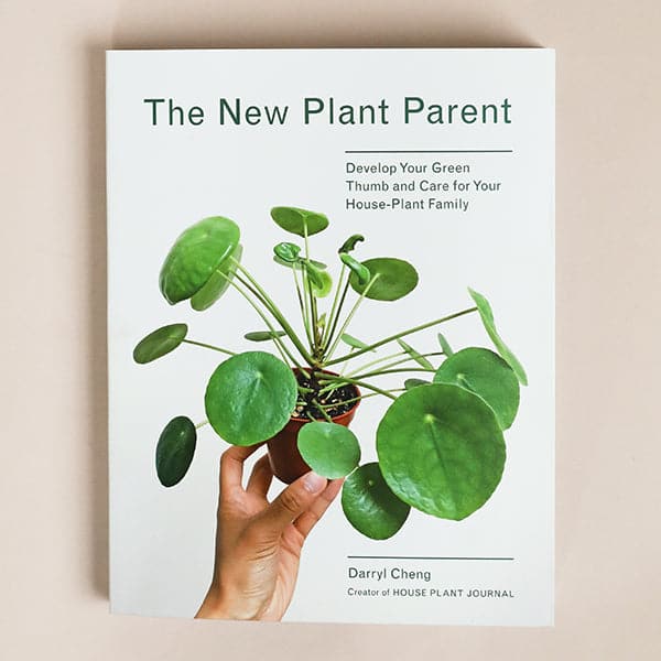 This book has a white cover reading &#39;The New Plant Parent&#39; across the top of the cover. A forest green Peperomioide plant with circular leaves and long stems is held in the center. 