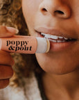 This is a close up picture of a woman’s mouth. She is holding a round peach tube up to her lips. On the left side of the tube is black text that reads ‘poppy & pout.’