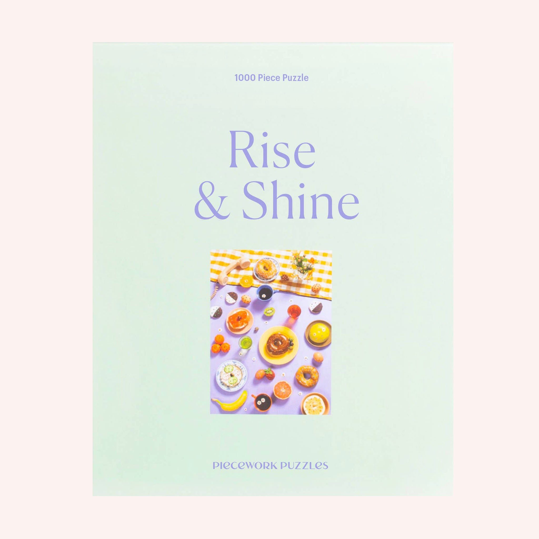 A mint green cardboard boxed puzzle with the words "Rise & Shine" in a light purple text along with an image of an array of bedazzled breakfast items including bagels, fruits, sunny side up eggs and more.