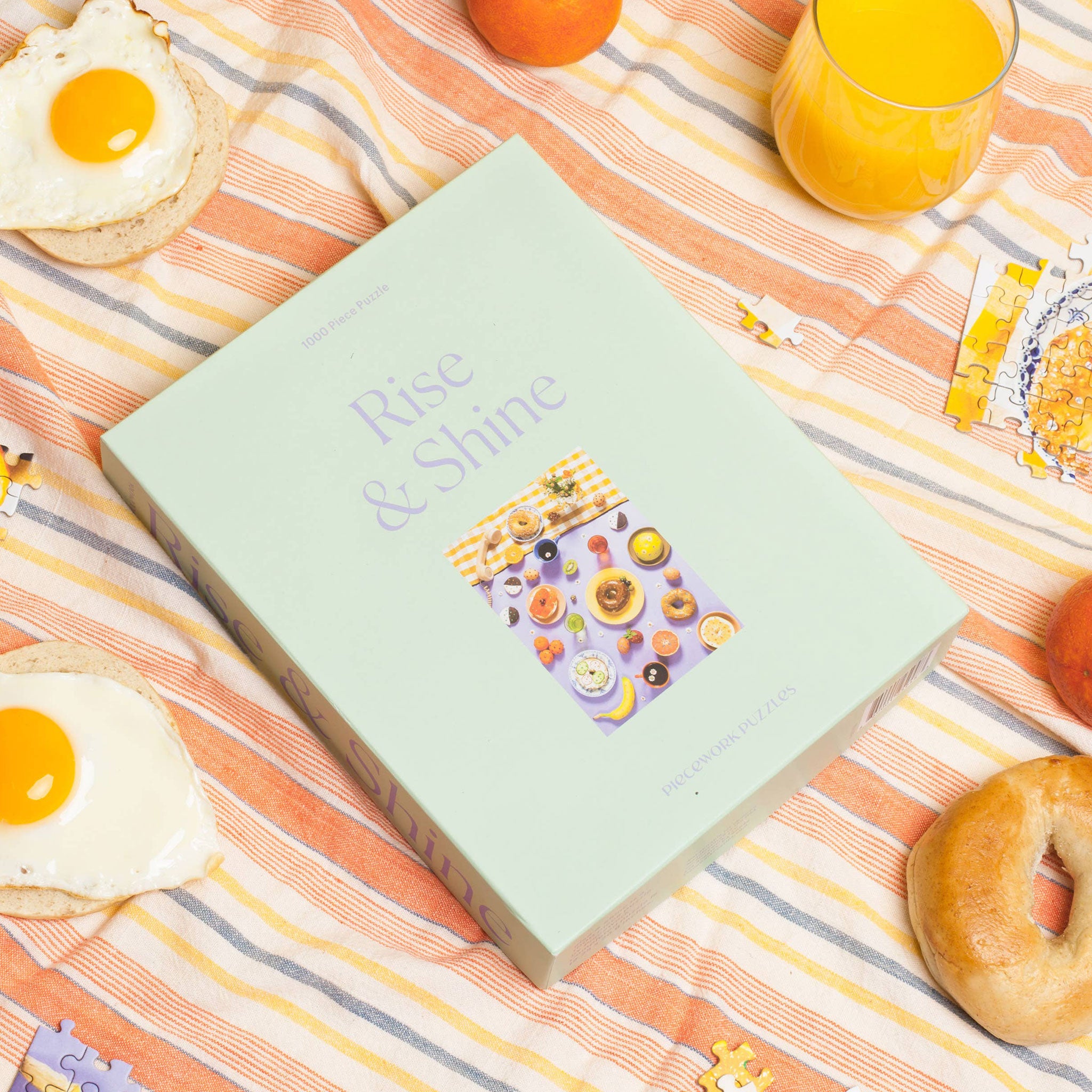 A mint green cardboard boxed puzzle with the words "Rise & Shine" in a light purple text along with an image of an array of bedazzled breakfast items including bagels, fruits, sunny side up eggs and more.