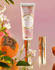 A tube of hand cream decorated with a pink floral pattern with gold text in the center that reads, "Voluspa Saijo Persimmon". The tube of hand cream has a gold pump and gold cap. It is photographed around florals and persimmon fruits.