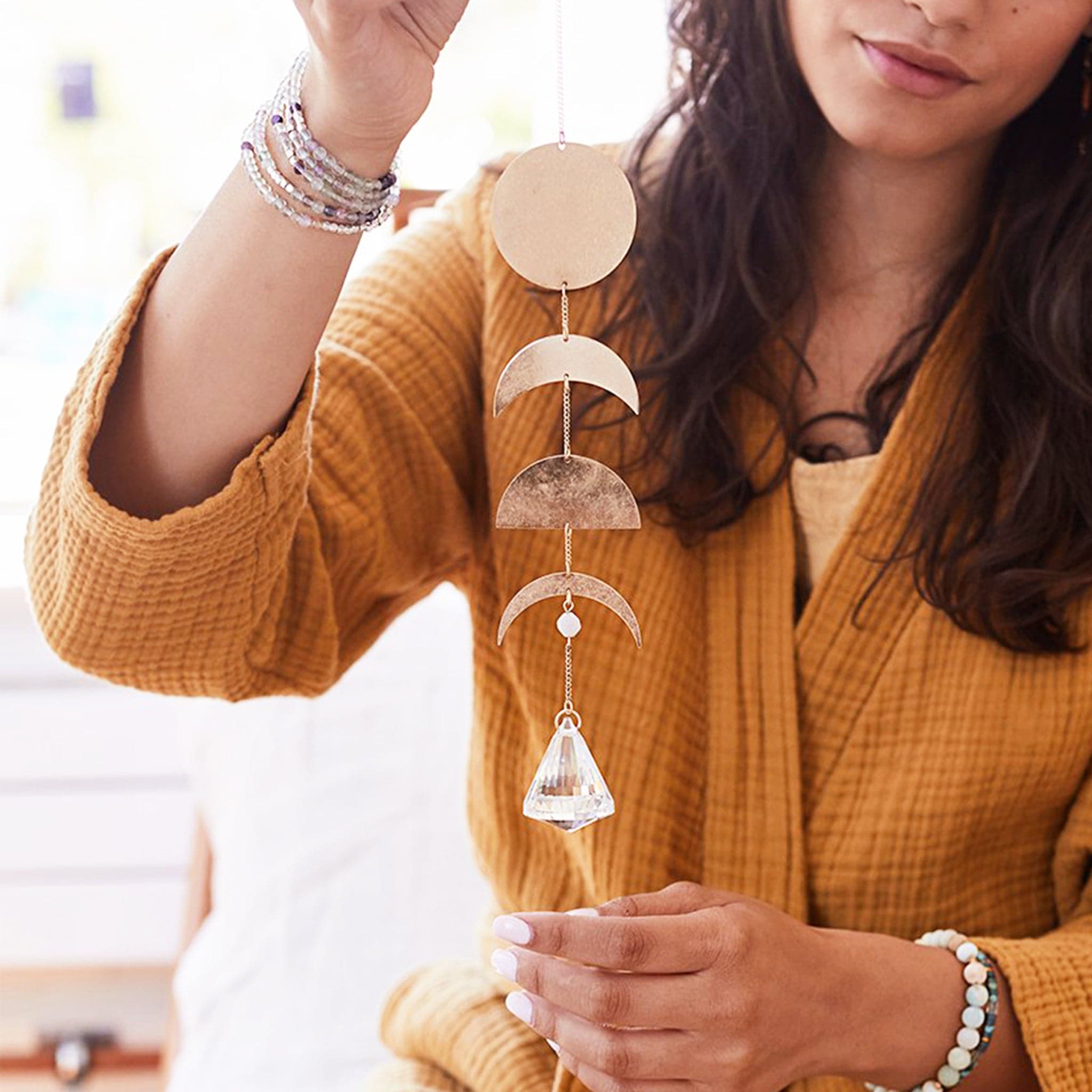 This is a picture of a woman holding wearing a burnt orange jacket. She has long brown hair. In her right hand she is holding a suncatcher. It is a gold chain with a clear, crystal prism at the bottom. Above that is a brown bead. Next is a gold, metal crescent moon shape. Next is a gold, metal half circle. Above that is another gold, metal crescent moon shape just a little thicker. At the top is a gold, metal circle. 
