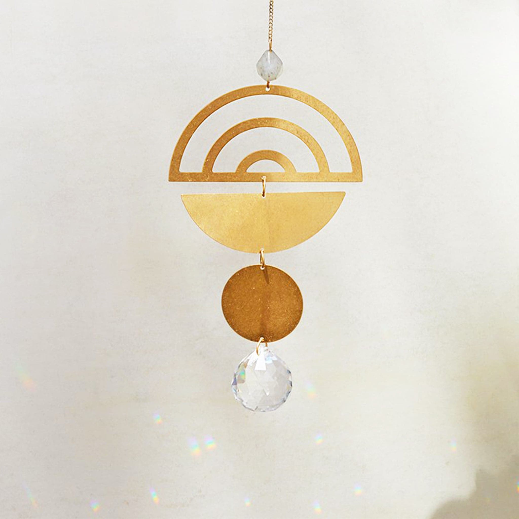 Hanging in front of a white wall is a light catcher. Starting at the top is a dark bead. Next is a gold, metal half circle with the arch at the top. There are three arch cut outs in the half circle. Attached by a small, gold hoop is an upside down, gold half circle. Attached by another gold hoop is a small gold circle. At the bottom is a circle, clear crystal. 
