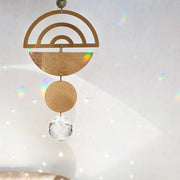 Hanging in front of a white wall is a light catcher. Starting at the top is a gold, metal half circle with the arch at the top. There are three arch cut outs in the half circle. Attached by a small, gold hoop is an upside down, gold half circle. Attached by another gold hoop is a small gold circle. At the bottom is a circle, clear crystal. 
