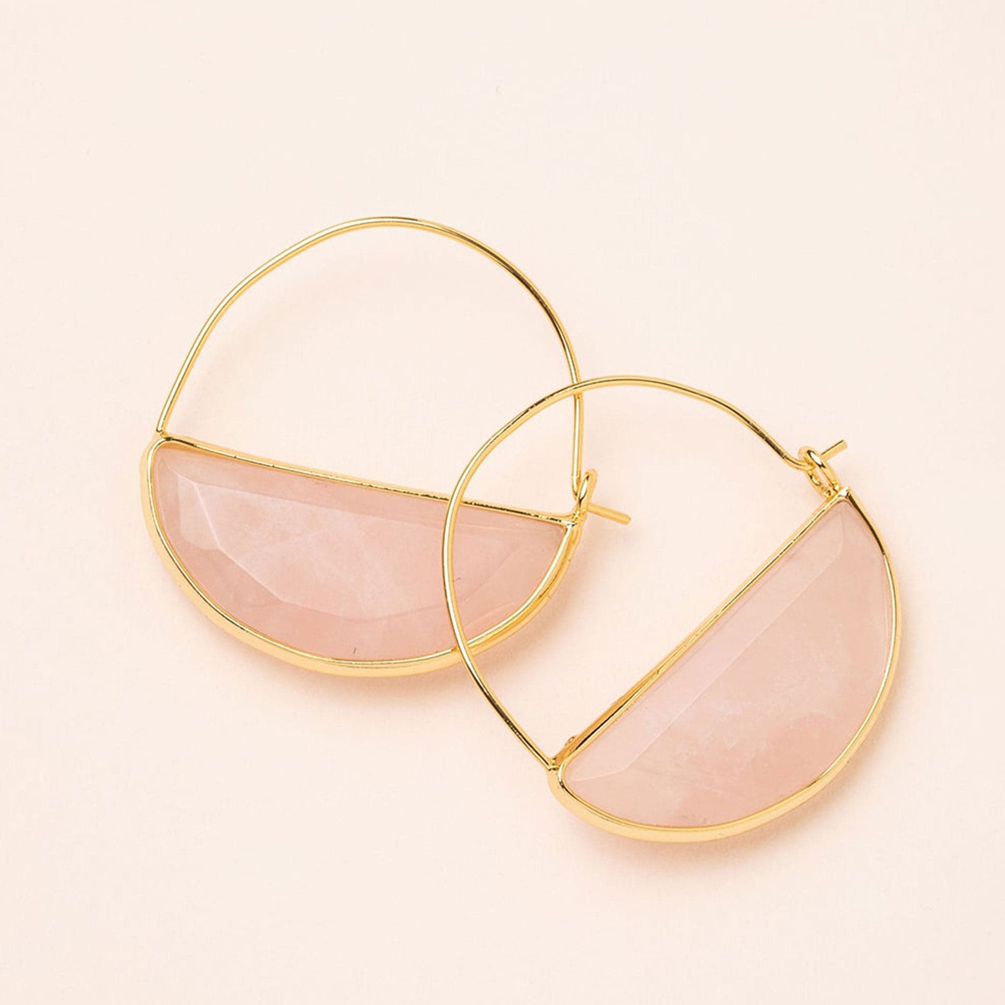 In front of a cream background is a pair of gold earrings. The top is a thin gold hoop. The bottom is a rose quartz half circle with a gold border. 
