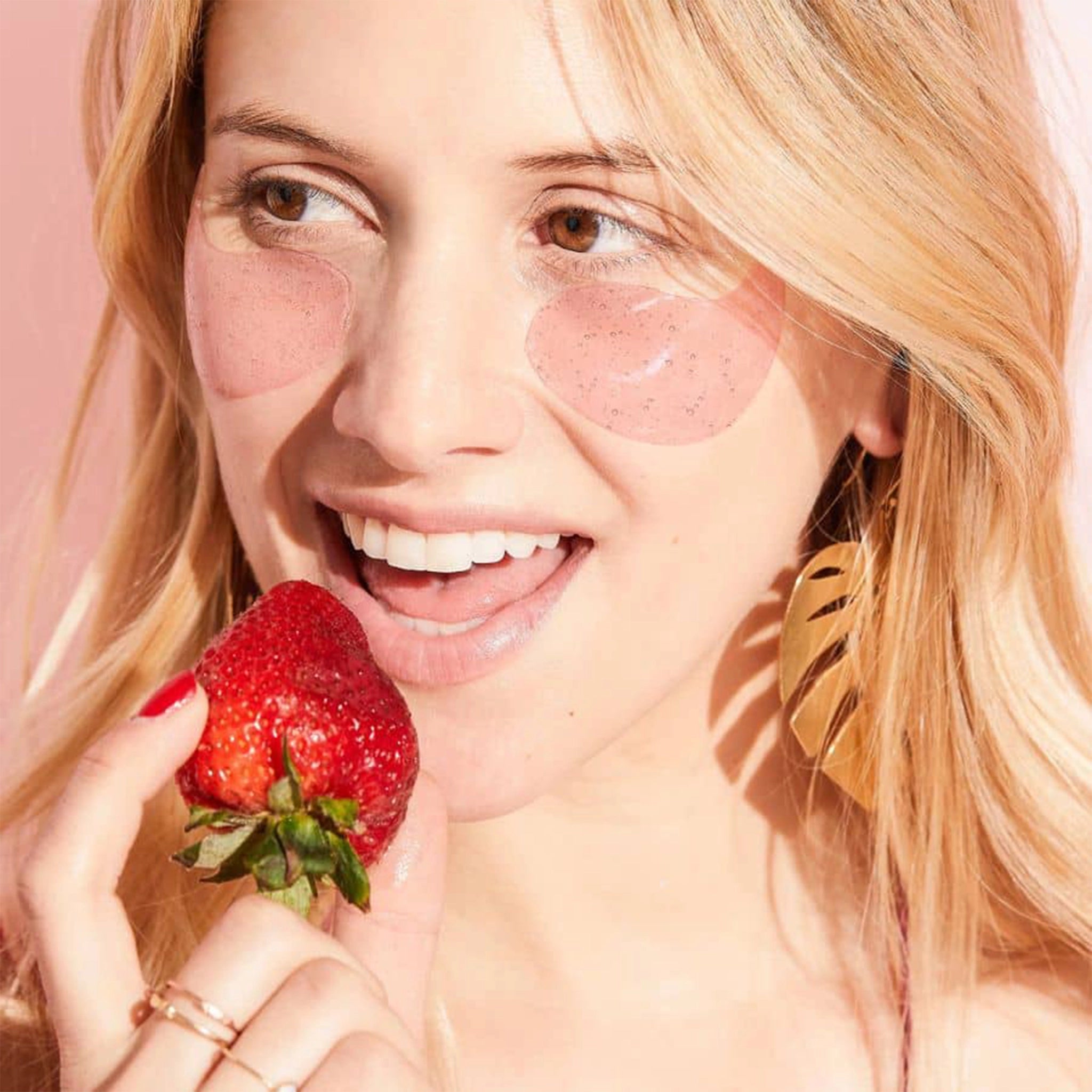 A model wearing the pink gel under eye masks while eating a strawberry.