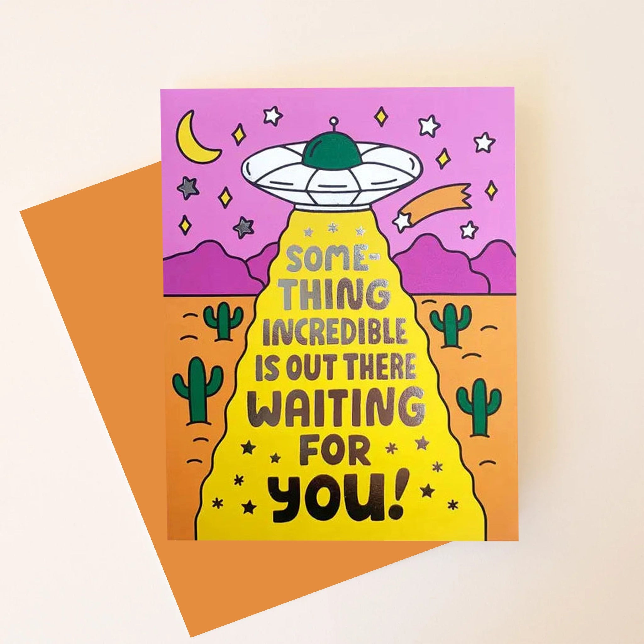 A vibrant greeting card  with an illustration of a spaceship floating in a desert, detailed with twinkling stars, green cacti, and a fuchsia mountain scape and reads, "Something incredible is out there waiting for you!". Also included is a coordinating orange envelope.