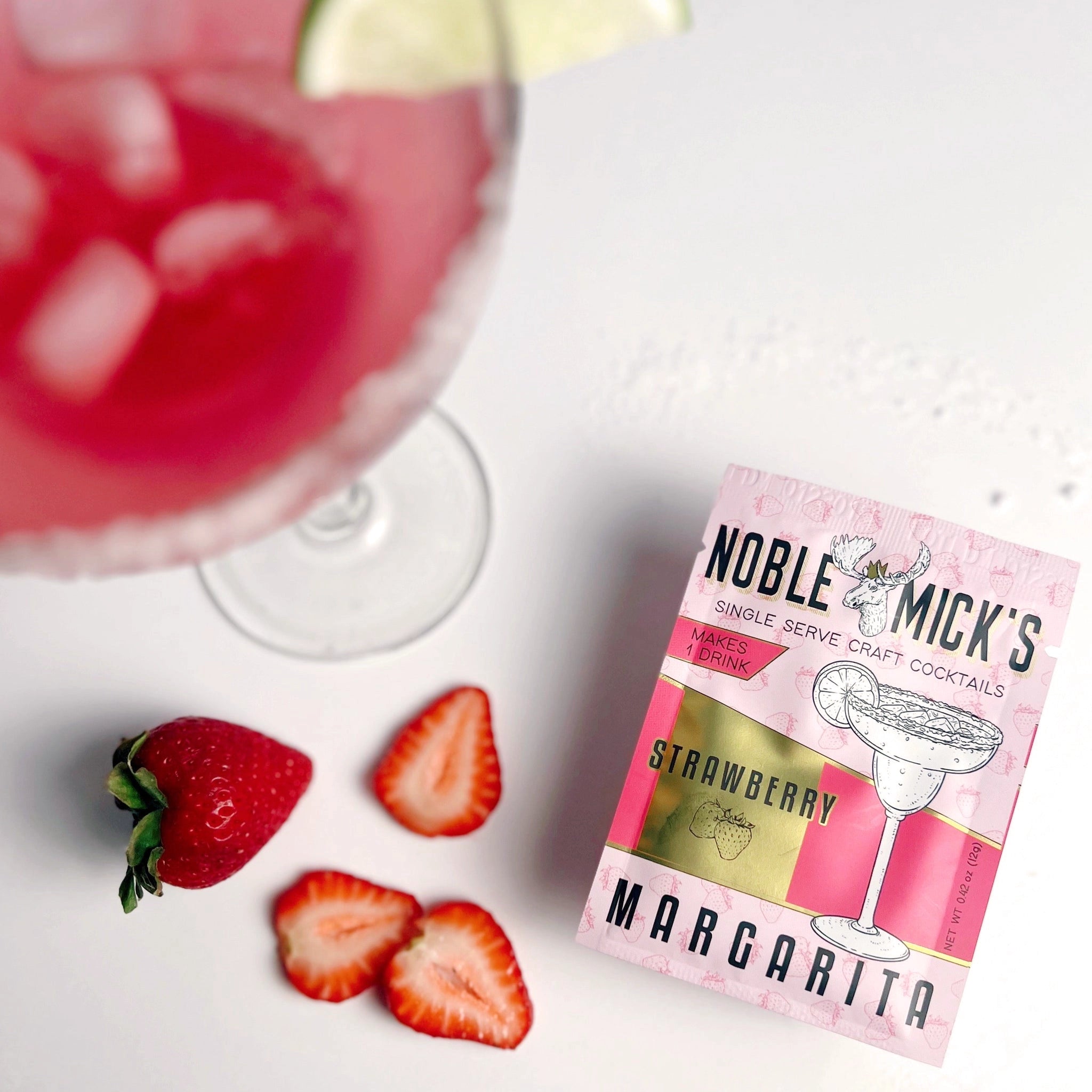 On a white background is a hot pink and light pink packet of cocktail mix that reads, "Noble Micks Single Serve Craft Cocktails Strawberry Margarita" photographed next to a strawberry and a few slices as well as a cocktail glass filled with a pink liquid. 