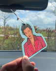 Light aqua air freshener with display the top half of Harry Styles wearing an iconic pink polo dot suit and cross necklace. Air freshener hangs on a white elastic loop from car mirror. A hand holds the air freshener in the light of the car window.