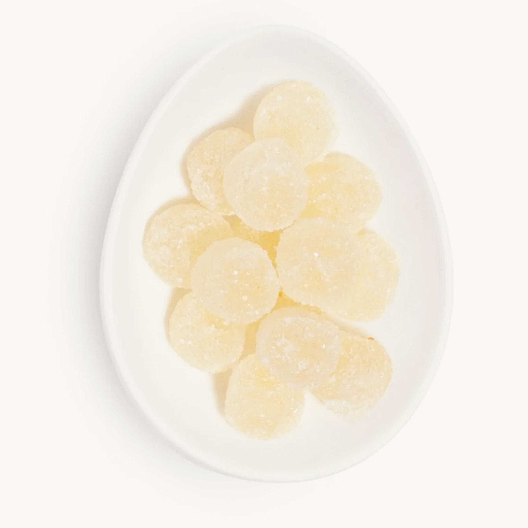 Birds eye view of a white egg shaped dish filled with light yellow candies. The candies are coated in white sugar. 