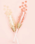 Birds eye view of a pink background. There are two champagne glasses that cross at the stem of the glass. The bottom of the glasses are covered with gold confetti. Spilling out of the class on the right is round light pink candies dusted with white sugar. On the left is round clear candies dusted with white sugar.