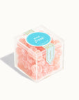 In front of a white background is a clear plastic cube filled with round light pink candies dusted with white sugar. On top is a vertical light blue sticker with gold circles on it. In the middle of the top is a bright blue circle sticker. Inside the circle is white text that reads ‘rosé frosé.’
