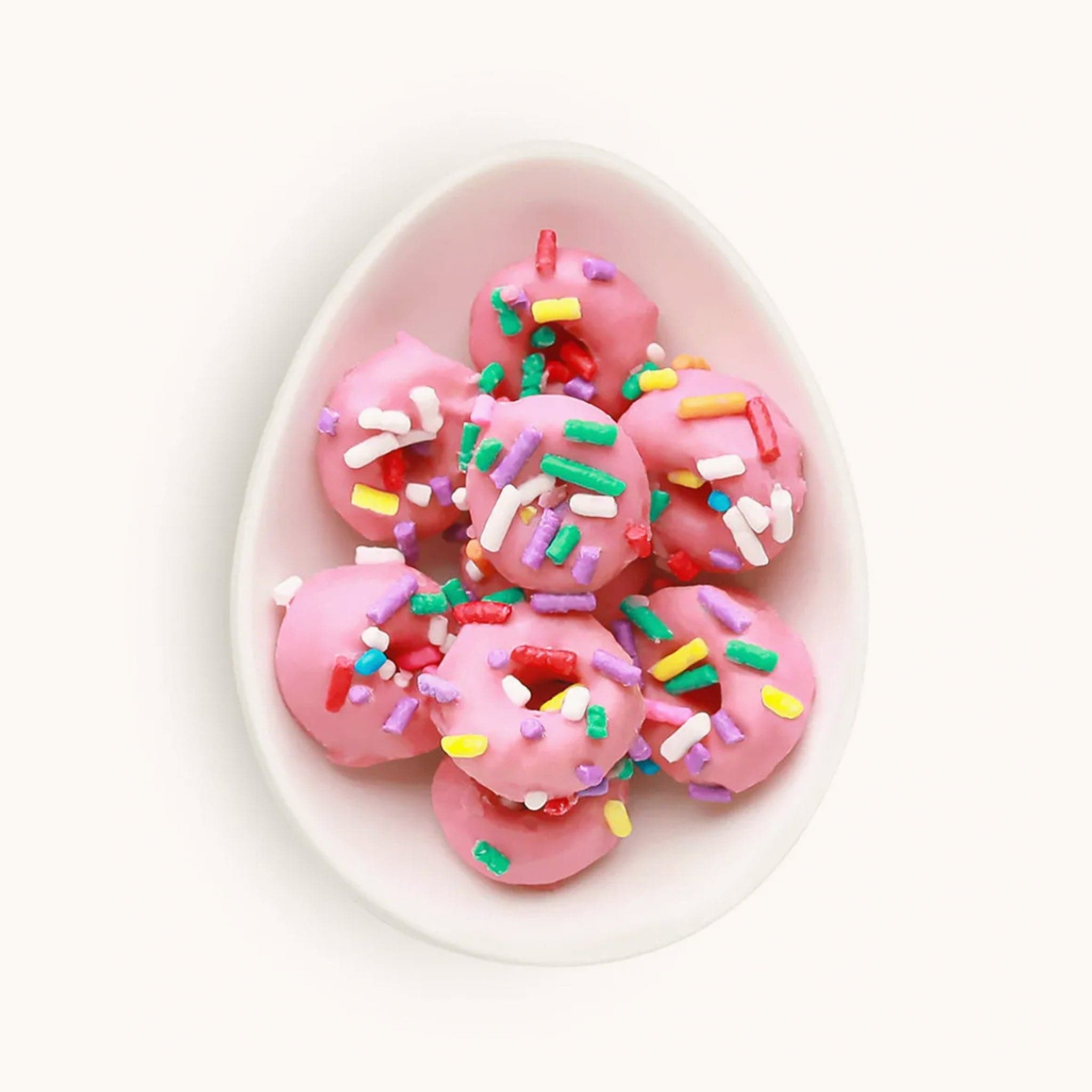 Miniature pink donut candies topped with colorful sprinkles. The candies are placed in a small white bowl. 
