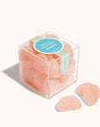 Plastic cube box filled with grapefruit gummies covered in sugar crystals. The clear box reads 'Tequila Grapefruit Sours' on a deep teal label. Three pieces are placed in front of the box.