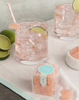 Plastic cube box of grapefruit gummies positioned on a soft pink cocktails garnished with gummies and limes. A small bowl filled with gummies is positioned next to the pair of glasses.