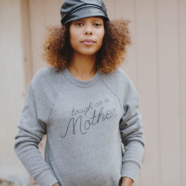 Soft grey screen-printed crewneck reading tough as a mother in black cursive. The crewneck is modeled by a woman with curly hair and black leather conductor hat. 