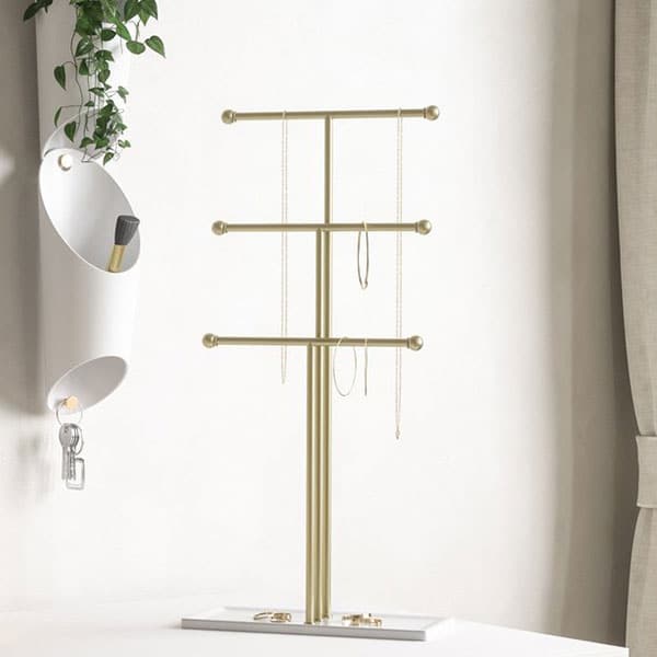 This gold, trigem three-tier jewelry stand stands tall with three plated bars and white base. A variety of necklaces and earrings are displayed on each tier. The stand is displayed next to a white wall-mounted key ring holder and the dangling green vines of a plant. 