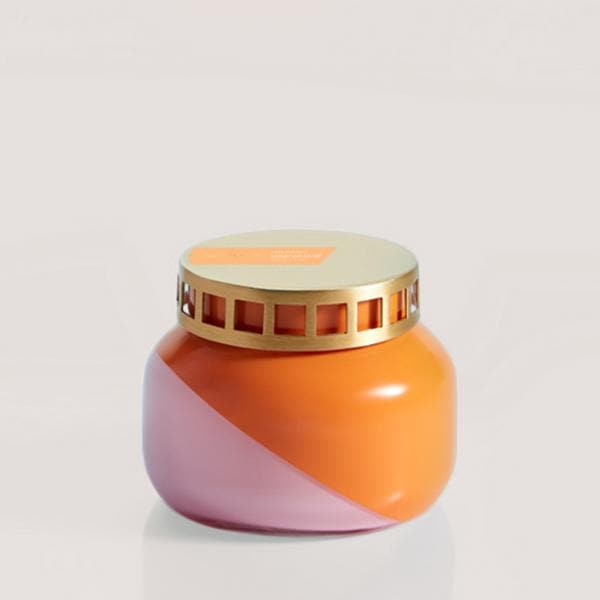 A squatty, round glass jar sits in front of a white background. The jar is split diagonally with the top half being a bright orange color and the bottom half being a soft pink color. The top of the jar is a lot narrower than the rest of the jar. The lid is a light gold color with one row of square cut outs around the side of the lid. There is an orange, rectangular shaped sticker on the left side of the lid. 