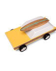 Birds eye view of wooden station wagon model with elongated daffodil yellow body with walnut veneer-paneled sides. Wheels are jet-black with white and silver rims. The windowed top half consists of a single white trapezoid shaped wooden block. A magnetic beech wood surfboard lays directly on top with a sea foam stripe a 'M' logo.