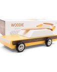 This wooden station wagon model has an elongated daffodil yellow body with walnut veneer-paneled sides. Wheels are jet black reading 'Candylab Mo-To' around white and silver rims. The windowed top half consists of a single white trapezoid shaped wooden block. A magnetic beech wood surfboard lays directly on top. A white packaging box is propped behind displaying 'Woodie' in orange lettering and a picture of the model itself.