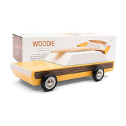 This wooden station wagon model has an elongated daffodil yellow body with walnut veneer-paneled sides. Wheels are jet black reading 'Candylab Mo-To' around white and silver rims. The windowed top half consists of a single white trapezoid shaped wooden block. A magnetic beech wood surfboard lays directly on top. A white packaging box is propped behind displaying 'Woodie' in orange lettering and a picture of the model itself.