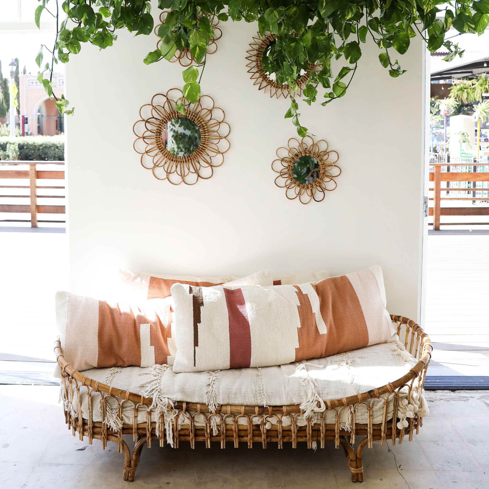 a rattan day bed with cream blanket and rust, cream, pink detailed large pillow sits in front of a white wall with rattan flower shaped mirrors and lush plants hanging from the ceiling.