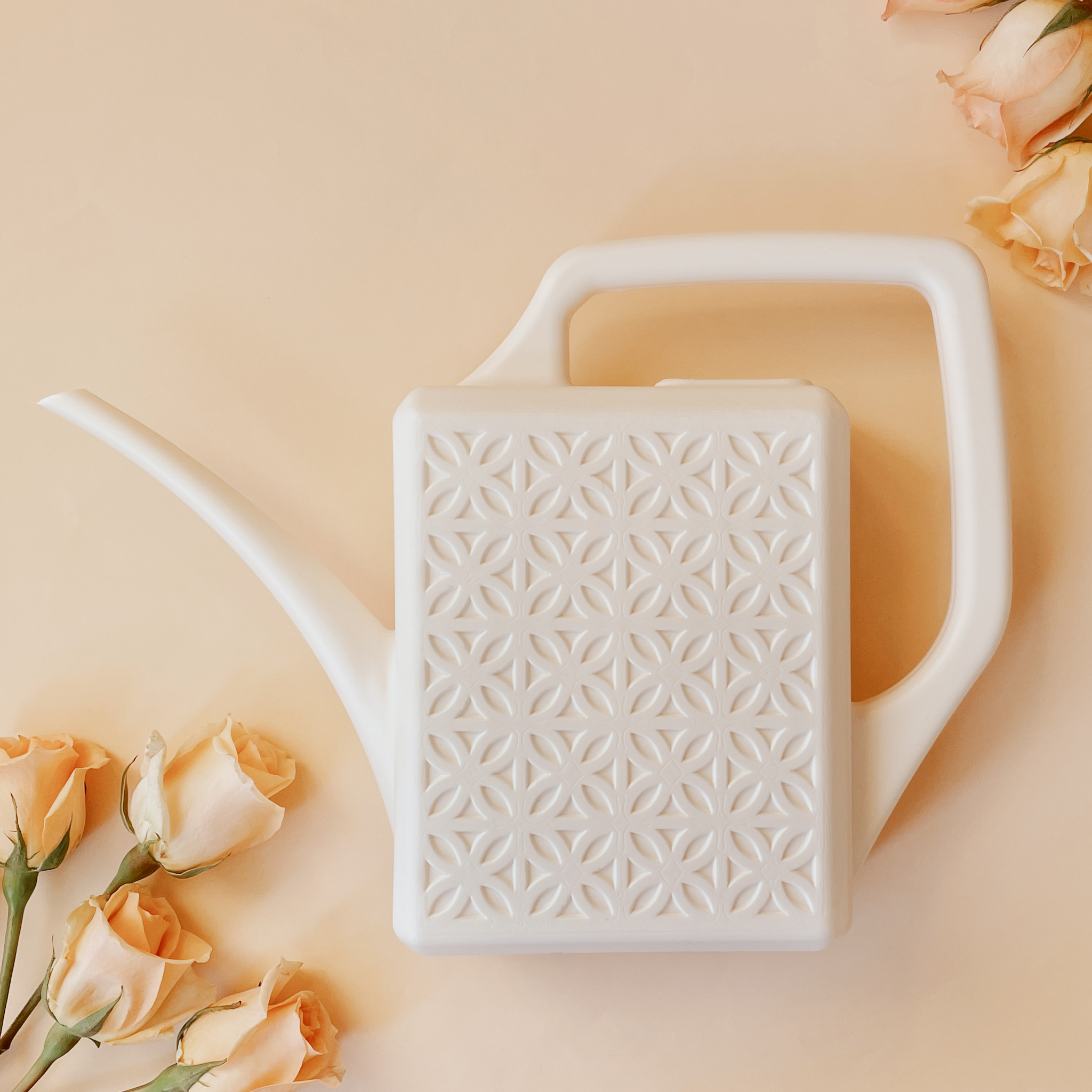 a white watering can with breeze block detail lies with peach colored roses