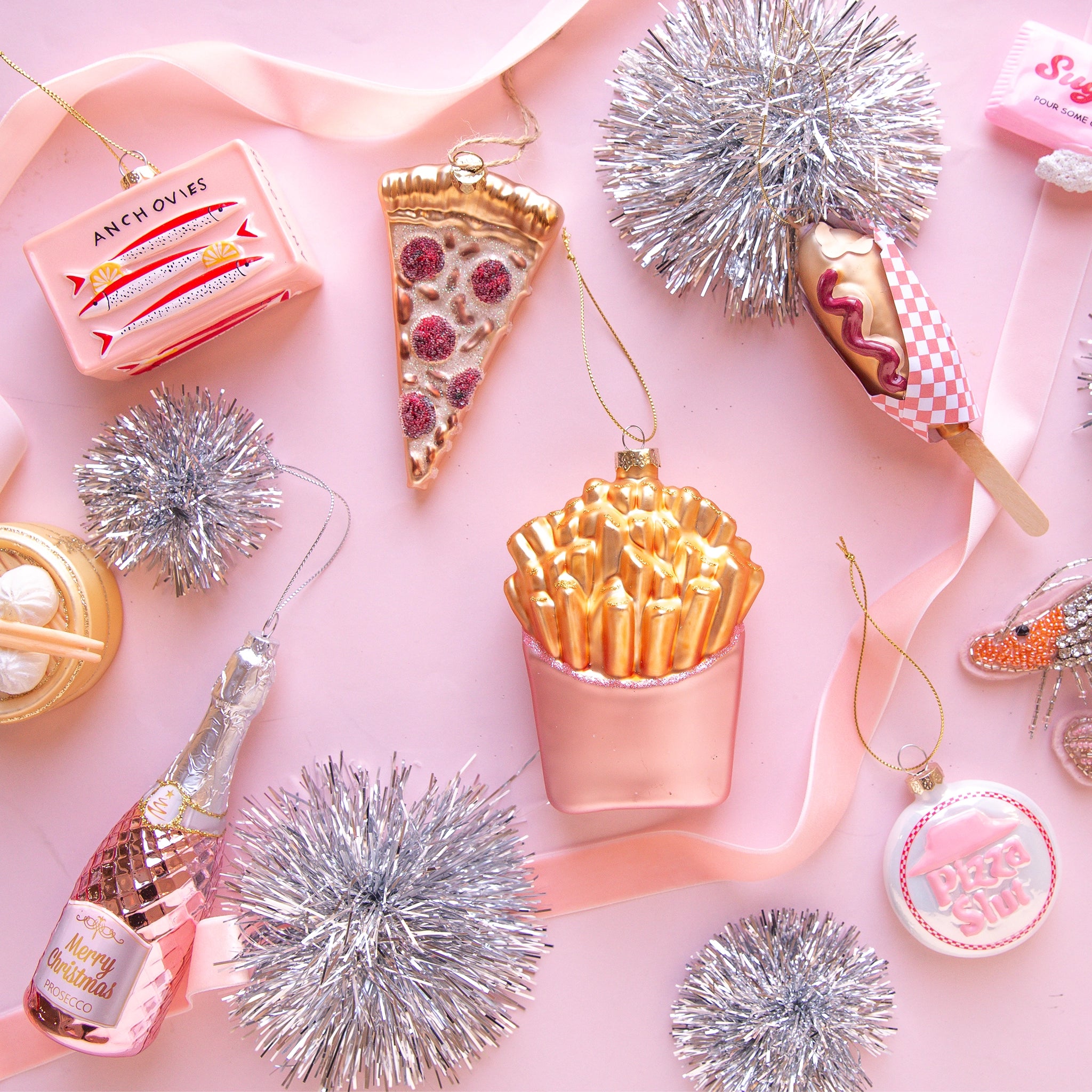 On a pink background is an assortment of glass ornaments in the shape of foods and staged next to pink ribbon and silver tinsel pom poms.