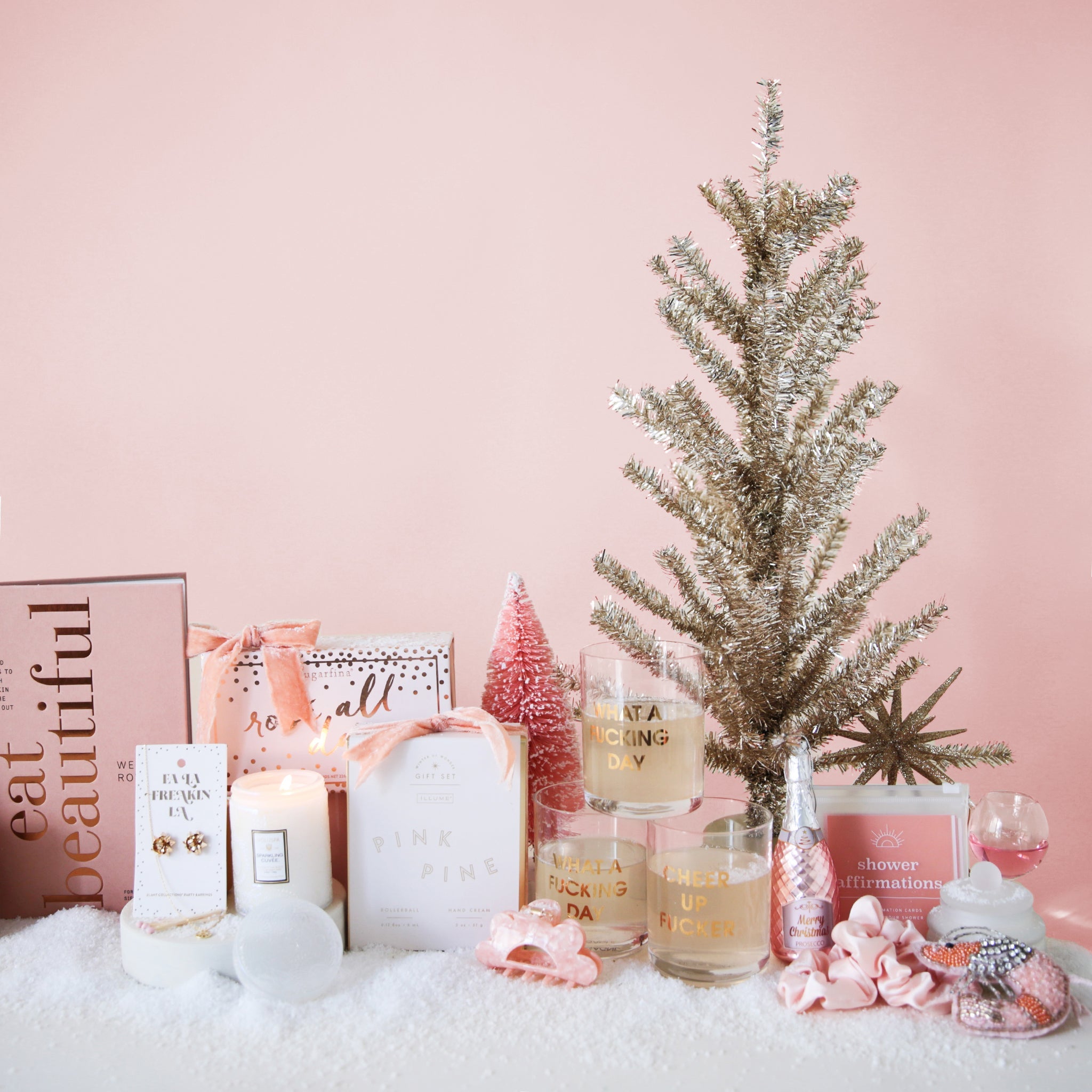 a pink book named eat beautiful, a pair of gold gift bow earrings, a white lit candle, a sugarfina rosé all day gift box, a pink hair clip, three drinking glasses that read what a f-ing day, pink ornaments, all on a pink ground with snow