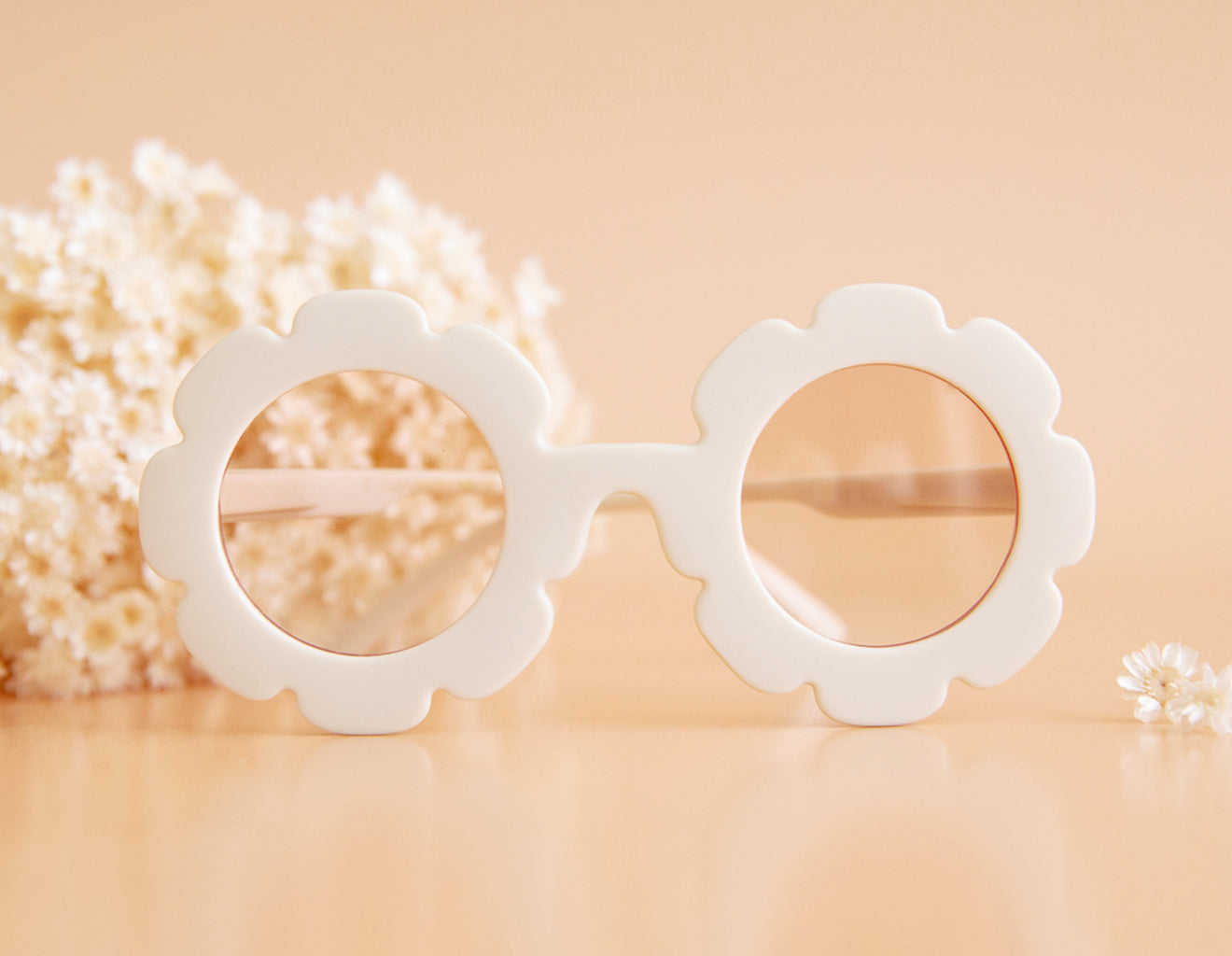 On a peachy background is a white flower shaped pair of sunglasses with a light pink lens, photographed next to ivory dried florals. 