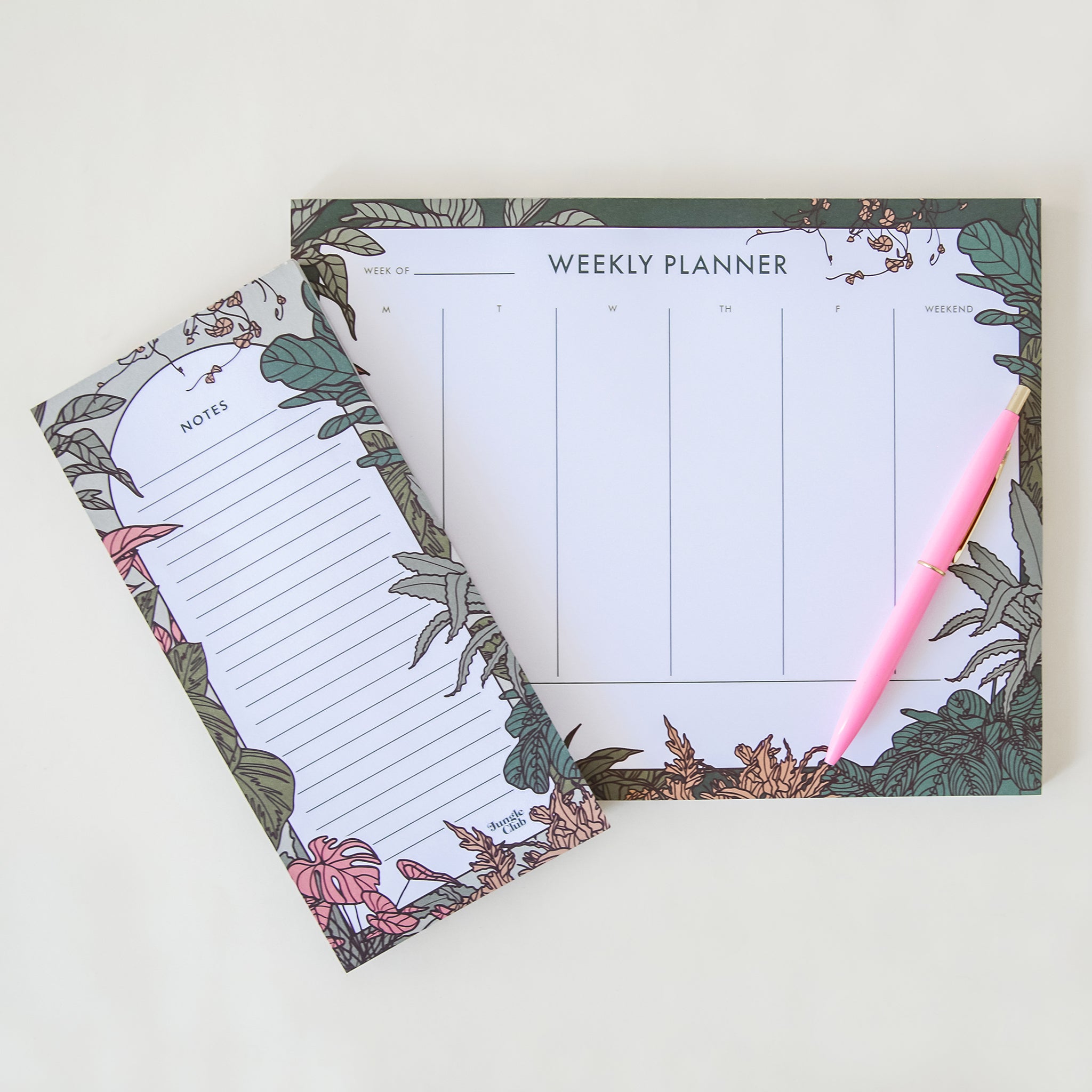 a notepad with pink and green plant illustrations lays with a weekly planner notepad with similar illustrations and a pink pen lays on top. all lay on a white ground. 