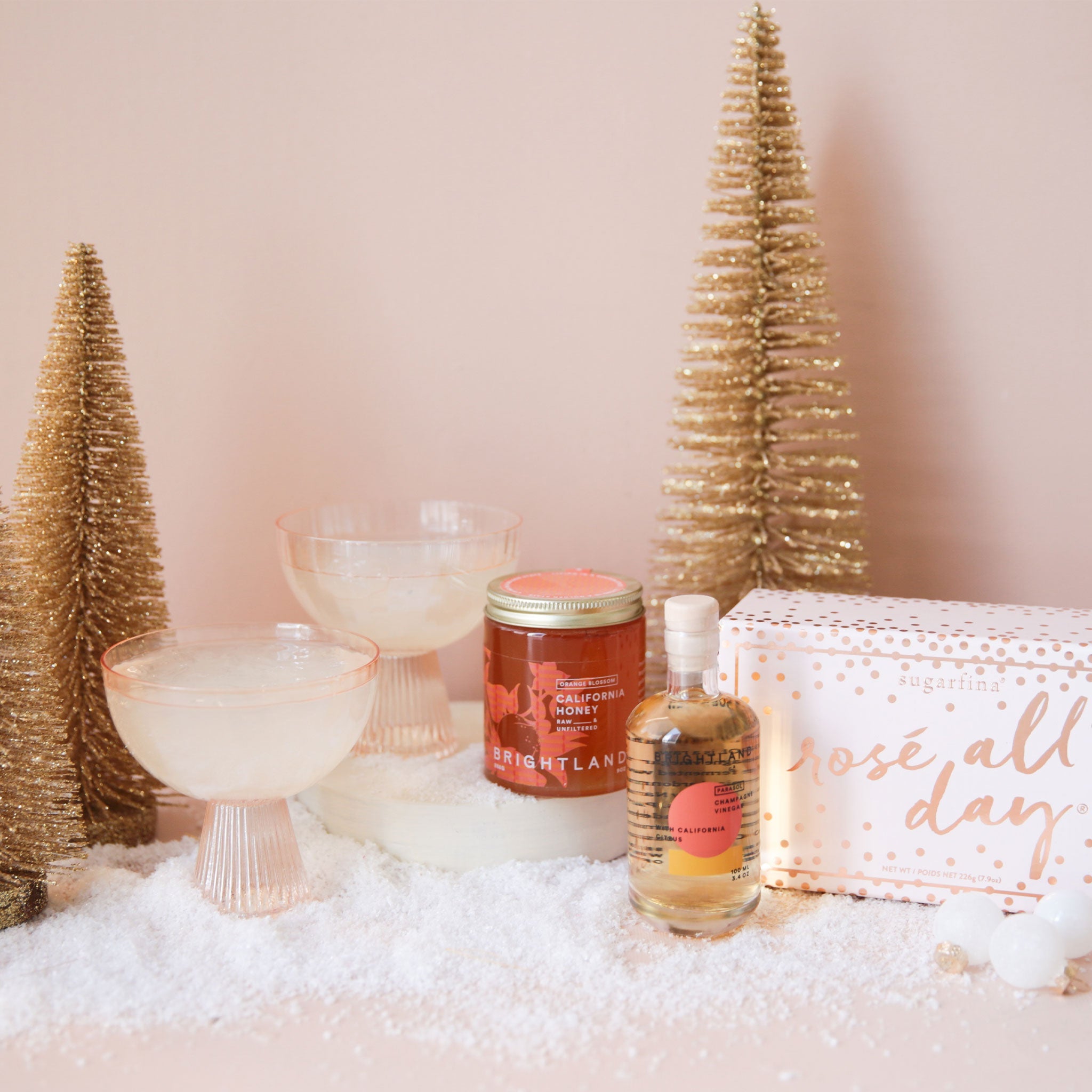 two coupe glasses filled with champagne, a small honey jar, a small jar of champagne vinegar, a pink rosé all day sugarfina gift box all on a soft pink ground with gold christmas trees and snow
