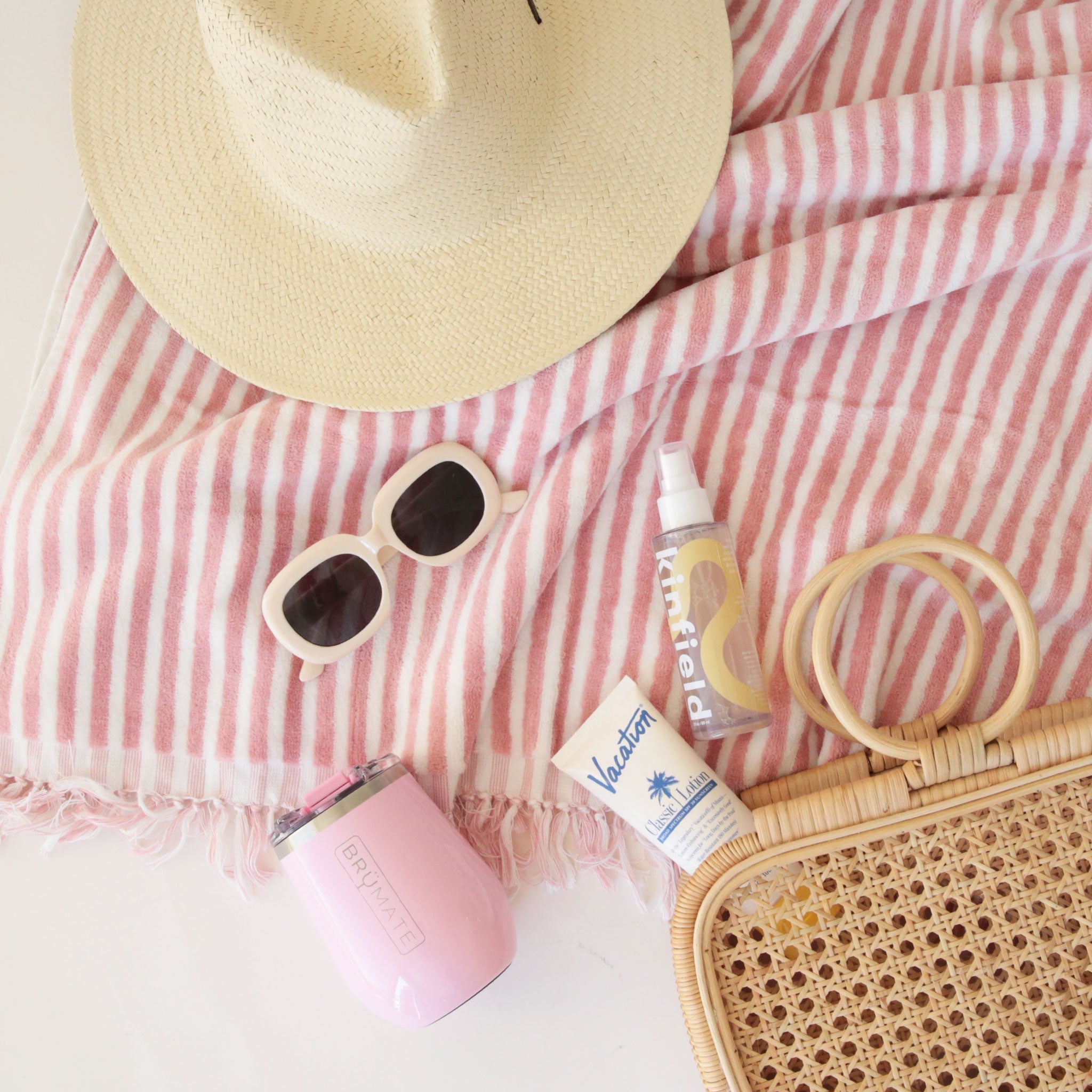 a woven tote, sunscreen, pink wine tumbler, beige vintage sunglasses and a straw hat sit on a white and pink striped beach towel