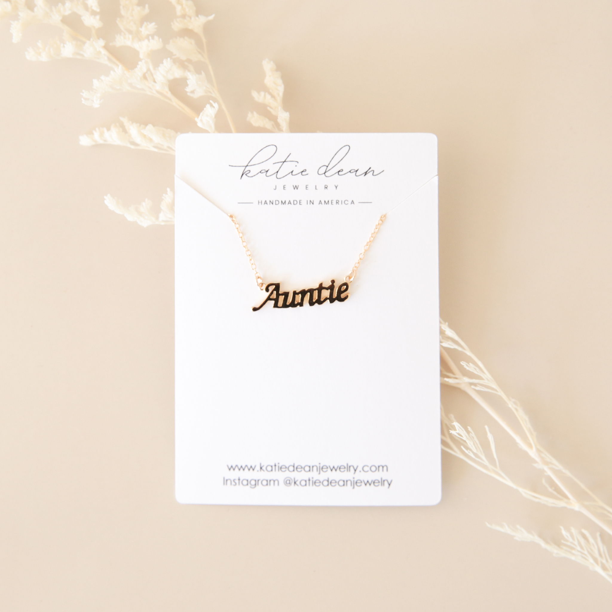 a gold necklace reads 'auntie' on a white jewelry card and sitting on a beige background with cream dried florals