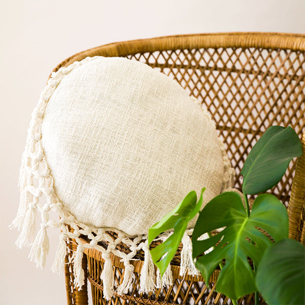 a cream, round tassel pillow sits on a rattan chair with a lush plant in the foreground