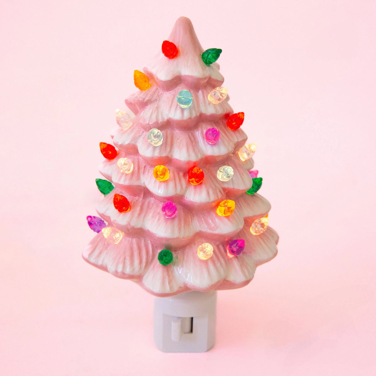 A pink Christmas tree-shaped night light with layered ruffled tiers, decorated with colorful lit bulbs in red, green, yellow, orange, purple, and white, mounted on a white plastic switch base against a solid pink background.