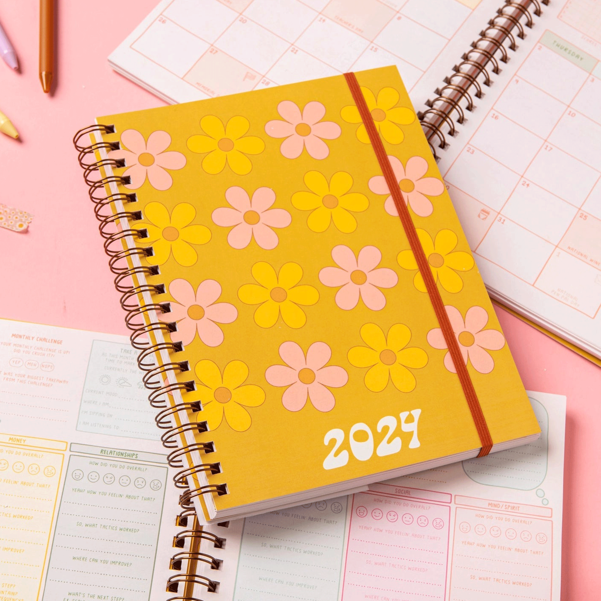 On a pink background is a yellow planner with a yellow and pink daisy daisy print with white text that reads, "2024". 